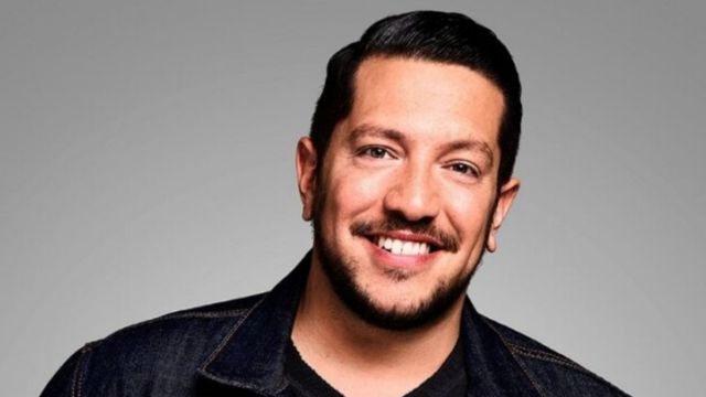Sal Vulcano wearing sky blue jacket and black t shirt under it and smiling and giving normal posse