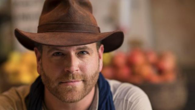 josh gates wearing brown hat and smiling posing for photo and blur flowers in backgrounds