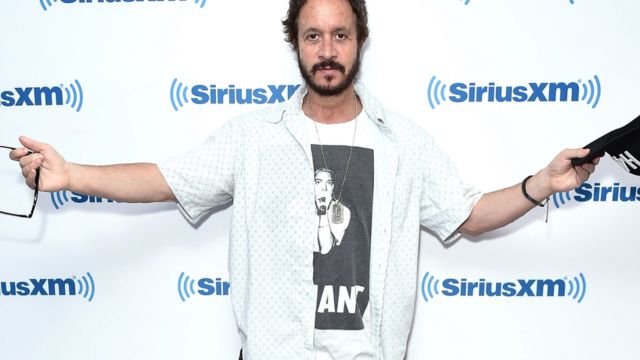 pauly shore wearing whoite t shirt and raising hands with one cap in left ahand and sunglass in right hand and with a bckground of sirius Xm